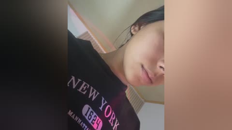 Video of a young woman with light skin, black hair, wearing a black T-shirt with \"New York\" in white letters. She's looking down, partially out of frame. Background shows a beige wall and a wooden ceiling.
