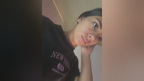 Video of a young woman with light brown skin and dark hair, wearing a black New York Yankees T-shirt, lying on her side, resting her head on her hand.