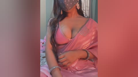 Media: Video of a South Asian woman with long dark hair, wearing a pink satin sari, a pink bra, and a face mask, sitting on a bed with pink and white pillows, in a dimly lit room.