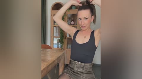 A video of a slender, fair-skinned woman with a ponytail, wearing a black tank top and high-waisted shorts, seated in a rustic kitchen with wooden shelves and greenery.