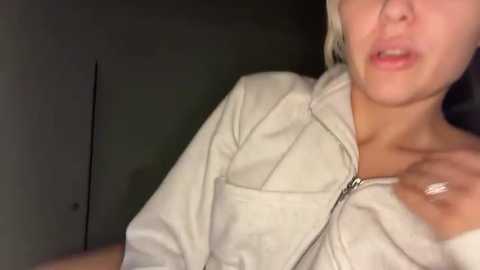 Video of a woman with short, platinum blonde hair, wearing a light gray hoodie, and a silver ring. Her lips are slightly parted, and she has a light skin tone. The background is dark and blurry, suggesting an indoor setting.