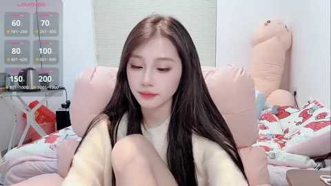 Media: Video of a young Asian woman with long black hair, fair skin, and a round face, sitting on a pink bed with a stuffed animal, wearing a beige sweater.