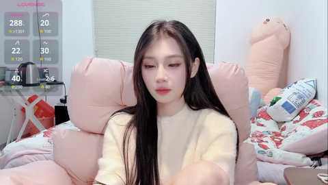 Media: A video of a young East Asian woman with long black hair, wearing a cream sweater, sitting on a pink bed with plush toys and a temperature gauge in the background.