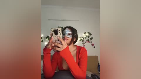 A video of a woman with medium skin tone, wearing a red cardigan, holding a phone with a white case, revealing a deep cleavage, and a glittery mask covering her eyes and nose.