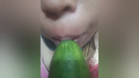 Video of a close-up view of a woman's face and a cucumber, with a blurry background. She has fair skin, a nose, and is wearing a pink shirt. The cucumber is partially inserted into her mouth.