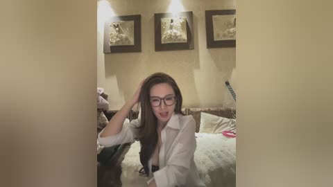 Video of a young woman with long brown hair, glasses, and a white shirt, seated on a bed in a dimly lit bedroom with beige walls and framed artwork.