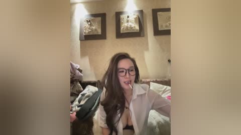 A video of a Caucasian woman with long brown hair, wearing glasses, and a white blouse, smoking a cigarette in a dimly-lit bedroom. The room has beige walls, framed pictures, and a bed with a green blanket.