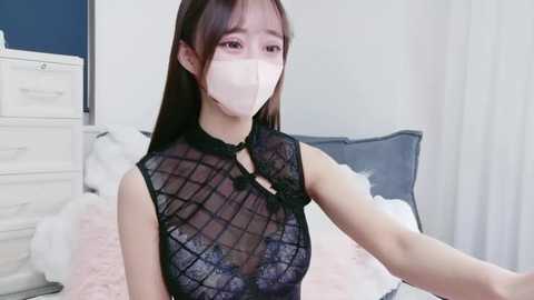 Video of an East Asian woman with long black hair, wearing a black, see-through lace dress, a face mask, and white gloves, sitting on a bed in a bright, minimalist room with white furniture.