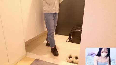 Video of a person wearing a light gray hoodie, blue jeans, and black shoes, standing in a beige-walled room. Next to them, a black bag and slippers are on the floor.