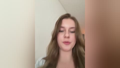 Video of a young, fair-skinned woman with long, wavy brown hair, smiling slightly, in a soft, blurred indoor setting with neutral tones.