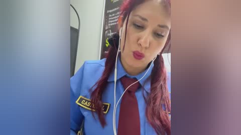 A video of a woman with long red hair, wearing a blue uniform with a name tag, listening to music through white earbuds. Background includes a blurred wall with a poster.