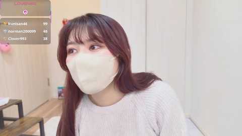 Video of a young Asian woman with long, straight brown hair, wearing a white face mask and a light sweater, sitting indoors against a white wall. Background includes a wooden table and a door.