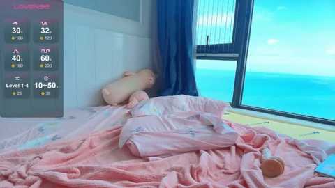 Video of a baby lying on a pink blanket in a bright room with large windows overlooking the ocean. The baby is wrapped in a light pink blanket, and there is a blue curtain beside the window.