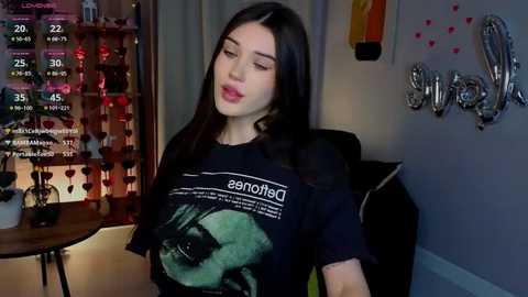 Video of a young woman with long black hair, wearing a black T-shirt with a green design, in a cozy room with a red curtain, silver wall decorations, and a wooden table.