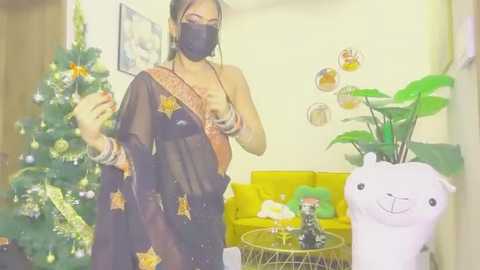 Video of a woman in a black saree with gold star patterns, wearing a face mask, standing next to a decorated Christmas tree and a yellow couch.