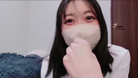 Video of an Asian woman with long black hair, wearing a white mask, white shirt, and blue jacket. She covers her mouth with her hand, in a room with a wooden door and blue fabric.