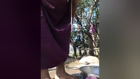 A video showing a person in a purple shirt and jeans bending over a metal bowl filled with pink and purple flowers in a sunny outdoor setting with trees and a fence in the background.