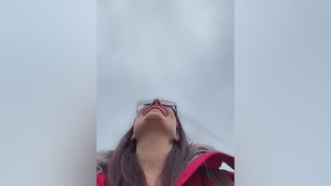 A video of a smiling woman with glasses, wearing a red jacket, taken from below, creating a dramatic upward angle. The background is a blurred, light-colored curtain.