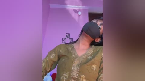 Media: Video of a person in a green, patterned blouse, wearing a black virtual reality headset, standing in a room with purple walls, framed pictures, and a ceiling light.