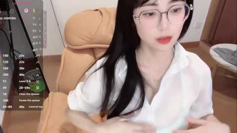 Video of an Asian woman with pale skin, long black hair, and glasses, wearing a white shirt, sitting on a beige chair in a room.