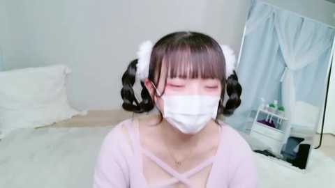 A video of an Asian woman with long black pigtails, wearing a white mask, pink top, and white headbands, in a light-colored bedroom with a white bed and blue curtains.