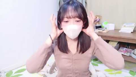 Video of an Asian woman with straight black hair, wearing a white face mask and beige button-up shirt, sitting on a bed with floral sheets, adjusting her hair in a bedroom with a wooden desk, green plant, and scattered items.