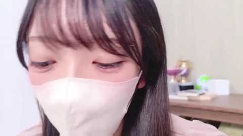 A video of an Asian woman with long, straight black hair and bangs, wearing a white surgical mask, standing indoors near a desk with office supplies and a phone.