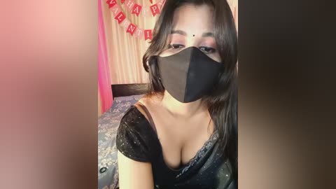 A video of a South Asian woman with medium skin tone, wearing a black face mask, black blouse, and a black saree, sitting on a bed with a pink and beige draped background.
