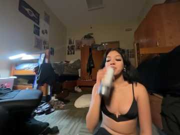 Video of a young woman with medium skin tone, dark hair, and medium breasts, wearing a black bra, sitting on a bed in a messy dorm room, eating a white snack.