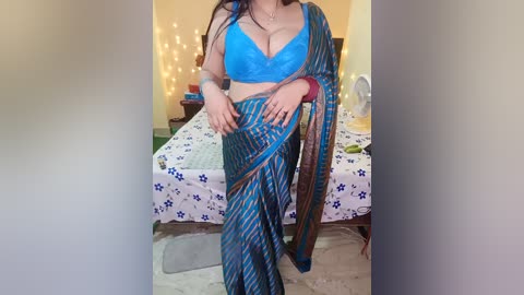 A video of a South Asian woman in a blue and gold sari, wearing a blue bralette, in a brightly lit bedroom with fairy lights, a bed with a floral duvet, and a fan.