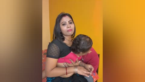 Video of a young woman with dark skin and long black hair, wearing a black top and red sari, cradling a baby in her lap. The background features a gradient of yellow and orange.