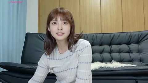 Video of an East Asian woman with straight, shoulder-length brown hair, fair skin, and a slender build, wearing a white, striped sweater, sitting on a black leather couch with a beige fur throw. Background features wooden-paneled walls and a blue curtain.