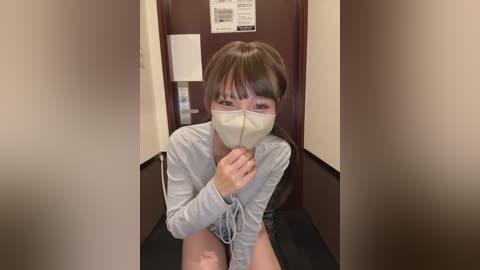 Video of an Asian woman with short brown hair, wearing a gray hoodie, white mask, and shorts, sitting in a narrow hallway with beige walls and a wooden door.
