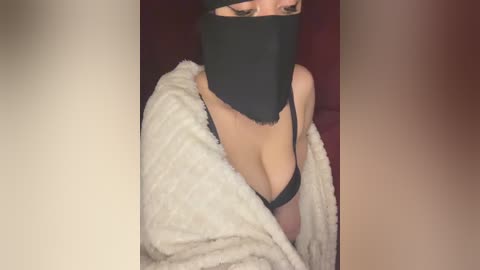 Video of a light-skinned woman with large breasts, wearing a black mask and black lingerie, covered by a white fuzzy blanket. The background is dark.