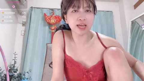 Video of an Asian woman with short dark hair in red lace lingerie, sitting on a chair with a blue curtain in the background, and a decorative red bird ornament.
