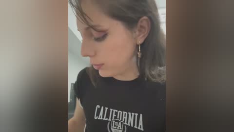 A video of a young woman with fair skin and brown hair, wearing a black t-shirt with \"CALIFORNIA\" in white text, and dangling earrings. The background is blurred, indoor setting.