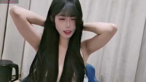A video of an East Asian woman with long black hair, wearing a sleeveless top, smiling while stretching her arms behind her head. She stands in front of a beige curtain in a minimalistic room.