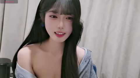 Video of an Asian woman with long black hair and pale skin, wearing a loose, off-shoulder denim shirt, smiling softly. Background features a beige curtain.
