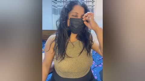 Video of a South Asian woman with long black hair, wearing a black face mask and a beige tank top, adjusting her mask, in a bedroom with a blue floral bedspread and a checkered wall tapestry.