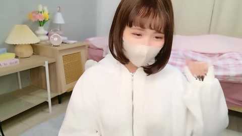 Video of a young Asian woman with straight brown hair and bangs, wearing a white face mask and a white hoodie, standing in a softly lit bedroom with a bed, nightstand, and lamp.
