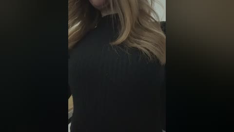 Video of a woman's face and shoulders in a dimly lit room, her long, wavy blonde hair partially covering her face. She wears a black turtleneck sweater. The background is blurred and indistinct.