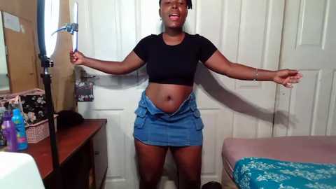 Video of a young Black woman with dark skin, medium build, and short hair, wearing a black crop top and denim mini skirt, posing with arms outstretched in a bedroom with white doors, wooden dresser, and bed.