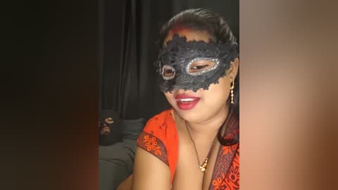 Video of a woman with medium skin tone, dark hair, and red lipstick, wearing a black lace mask with intricate patterns, an orange and black floral-patterned top, and a gold necklace. She is smiling.