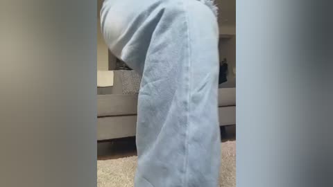 Video of a person bending over, wearing light blue jeans with a soft texture, standing in a carpeted living room with beige couch and white lamp.