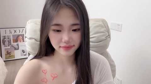 Video of a young East Asian woman with long black hair, fair skin, and a small heart tattoo on her left shoulder. She is seated in a beige chair, wearing a white top, in a softly lit room with a magazine on a table in the background.