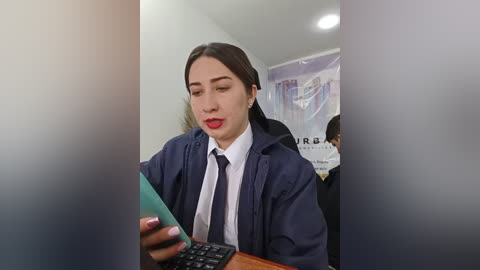 Video of a Latina woman with medium skin tone and long dark hair, wearing a navy-blue academic robe, white shirt, and tie, intently looking at a teal smartphone, seated in a blurred room with a whiteboard.