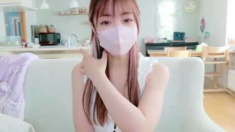 Video of an Asian woman with long, straight hair in a white sleeveless top and pink face mask, sitting on a light-colored sofa in a modern, bright kitchen with white walls, wooden furniture, and a clock on the wall.