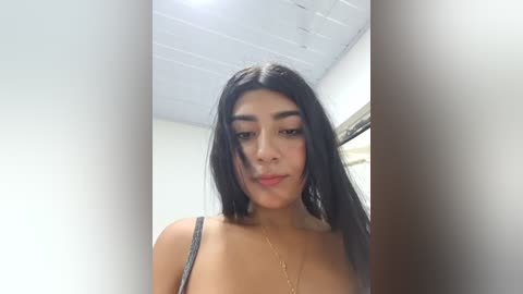 Video of a young Latina woman with long, straight black hair and medium skin tone, wearing a gray spaghetti-strap top, standing indoors with white ceiling tiles and a window in the background.