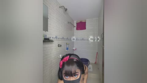 Video of a small, white-tiled bathroom with a black-haired woman in a pink headband, wearing a black mask, and a blue top, sitting on the toilet.