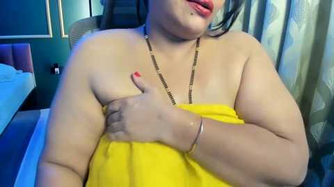 Video of a plus-sized woman with medium skin tone, dark hair, and full lips, partially covered by a yellow towel, in a modern bedroom with a bed and patterned curtains.